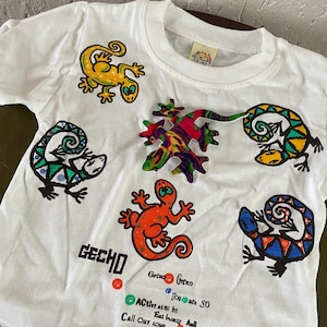 Puede incluir: Camiseta blanca con un diseño de gecko colorido. La camiseta tiene la palabra "Gecko" impresa en la parte delantera, junto con el texto "Gecko, Joy, Fun, SO Active, Eat healthy And Call Out loud, Wish DELIGHT".