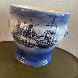 Könnte beinhalten: Ein blau-weißer Keramik-Pflanztopf mit einer holländischen Landschaftsszene mit einer Windmühle und einem Segelboot. Der Pflanztopf hat einen breiten, gebogenen Rand und zwei kleine Griffe. Der Sockel ist in einem dunkleren Blauton gehalten.