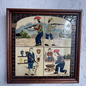 Könnte beinhalten: Ein gerahmtes Kunstwerk mit vier Keramikfliesen, die Jagdszenen zeigen. Die Fliesen zeigen Figuren mit Gewehren, einen Hund und eine Kochszene, alles vor dem Hintergrund von Landschaften und architektonischen Elementen. Der Rahmen ist dunkelbraun.