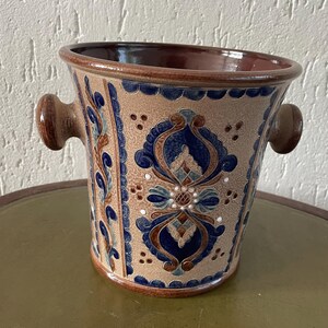 May include: A ceramic vase with a beige background and brown rim, featuring blue and brown floral patterns. The vase has two brown handles and sits on a green table. The design includes symmetrical floral motifs and decorative borders.