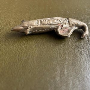 Op de afbeelding: Een kleine, antiek ogende metalen figuur van een olifant met een gekrulde slurf. De olifant heeft een gedempte bronzen kleur en geëtste details. Het beeldje ligt op een getextureerd, olijfgroen oppervlak.