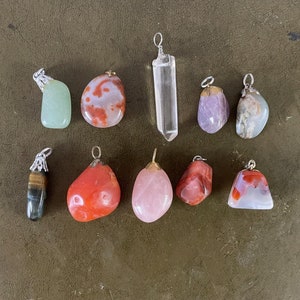 May include: A collection of ten gemstone pendants with silver or gold-colored metal loops. The stones vary in shape, color, and texture, including clear quartz, rose quartz, and other polished stones. The pendants are arranged on a dark green surface.
