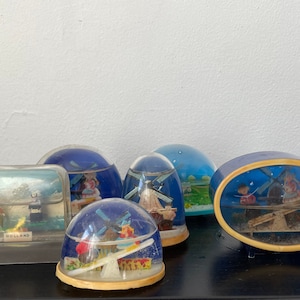 Può includere: Una collezione di sei sfere di neve in vetro vintage con diverse scene che raffigurano mulini a vento e figure olandesi. Le sfere di neve sono su una superficie scura. Una sfera di neve ha la scritta "HOLLAND" stampata sulla base.