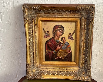 Op de afbeelding: Een ingelijst religieus icoon dat de Maagd Maria afbeeldt die baby Jezus vasthoudt. Het icoon is geschilderd in een traditionele stijl met gouden accenten en bevat de tekst "MP OY ПРОСТА СИЛ" in het Cyrillische schrift.