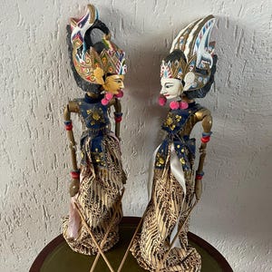 Puede incluir: Dos marionetas indonesias Wayang Golek. Las marionetas tienen tocados ornamentados, caras pintadas y trajes detallados en azul, dorado y tela estampada. Se encuentran sobre una superficie redonda y verde.