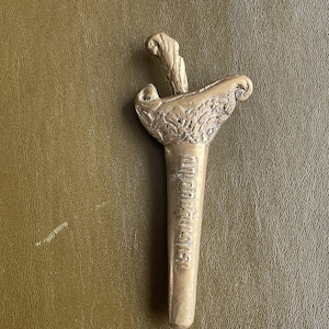 Op de afbeelding: Een gedetailleerde, antieke bronzen dolk met sierlijke gravures. Het heft heeft een gebogen ontwerp en het lemmet is voorzien van tekst. De dolk ligt op een getextureerd, olijfgroen oppervlak.