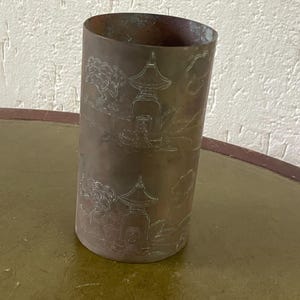 Può includere: Un contenitore cilindrico color bronzo con un disegno di paesaggio asiatico inciso. Il disegno comprende pagode e alberi. Il contenitore è leggermente affusolato e poggia su una superficie verde e marrone.