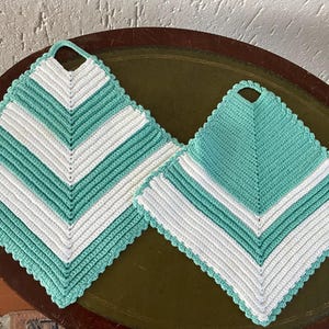 Könnte beinhalten: Zwei gehäkelte Topflappen im Zickzackmuster. Der größere Topflappen ist weiß und türkis, während der kleinere Topflappen türkis und weiß ist.