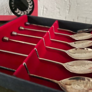 May include: A set of six silver spoons with ornate handles, displayed in a red velvet-lined box. The spoons are arranged in rows, with the handles facing up.