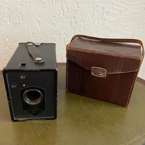 Puede incluir: Una cámara de caja negra vintage con un asa de transporte de cuero y una funda de cuero marrón. La cámara tiene una lente y varios diales. La funda tiene un cierre metálico.