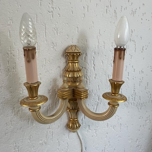 May include: A white and gold wall sconce with two candle-shaped light bulbs. The sconce has a decorative, ornate design.