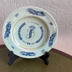 May include: A blue and white ceramic plate with a gold rim and a blue and white floral design. The center of the plate features a gold crown with the letter "B" inside, surrounded by a wreath of leaves. The text "7 Januari 1937 1962" is printed below the crown.