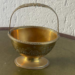 Könnte beinhalten: Kleiner, antiker Messingkorb mit Henkel. Der Korb hat eine runde Schalenform mit einem dekorativen Blumenmuster, das auf die Seite geätzt ist. Der Griff ist gewölbt und der Korb steht auf einem kleinen Sockel.