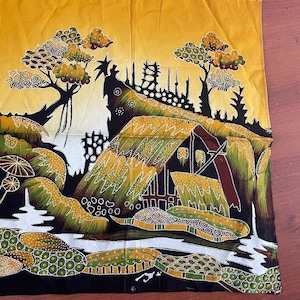 Op de afbeelding: Een batik-stofpaneel met een geel, groen en bruin ontwerp. Het ontwerp toont een gestileerd huis met een rieten dak, bomen en een rivier. De stof is versierd met kleine witte stippen.