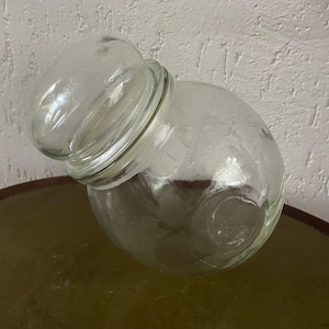 Op de afbeelding: Een heldere glazen pot met een afgeronde body en een deksel. De pot is gekanteld en onthult de inhoud. De pot staat op een rond, donkergekleurd oppervlak. De achtergrond is een getextureerde witte muur.