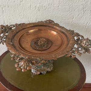 May include: An ornate copper serving dish with a decorative base and intricate details. The dish has a round, shallow bowl with a raised center piece.