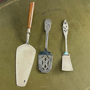 Puede incluir: Tres utensilios antiguos para servir sobre una superficie verde. Los utensilios incluyen una espátula para pasteles con mango de madera, una espátula para tartas con un recorte decorativo y una pequeña espátula con un diseño floral.