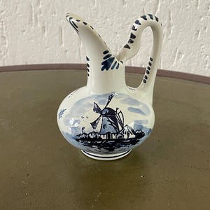 Op de afbeelding: Een kleine witte keramische kan met een blauw-wit windmolenontwerp. De kan heeft een handvat en een tuit.