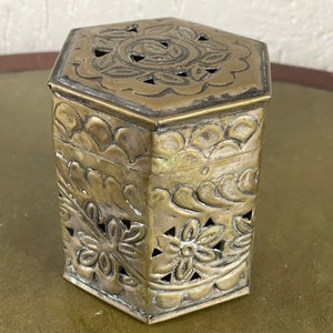 Puede incluir: Una caja hexagonal de latón con intrincados diseños florales y patrones recortados. La caja tiene una tapa y está hecha de latón brillante y envejecido.