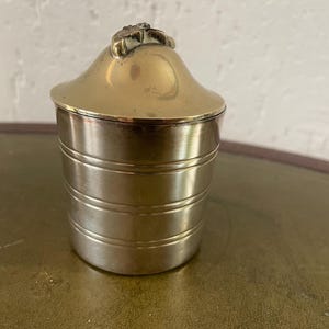 Könnte beinhalten: Ein goldfarbener Metallbehälter mit einem dekorativen Deckel. Der Deckel hat ein erhabenes Blumendesign.