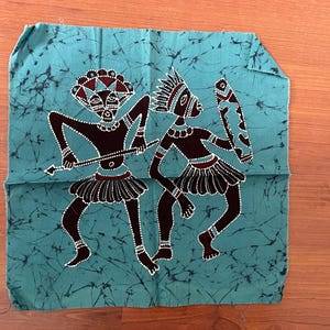 Könnte beinhalten: Ein türkisfarbenes Stoffquadrat mit einem Batik-Design, das zwei stilisierte Figuren in Schwarz mit weißen Punktakzenten zeigt. Eine Figur hält einen Speer, die andere einen Schild. Die Figuren tragen Kopfbedeckungen und Röcke.