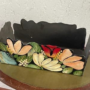 Peut inclure: Une boîte en bois noire avec un motif floral. La boîte présente des fleurs peintes à la main dans des tons pêche, rouges et jaunes, avec des feuilles vertes. La boîte est posée sur une table ronde avec une surface verte.