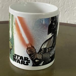 Op de afbeelding: Witte keramische mok met een Star Wars-thema. Het ontwerp toont Darth Vader en een lichtzwaard met een groene en rode gloed. De tekst "Star Wars" is op de mok gedrukt.