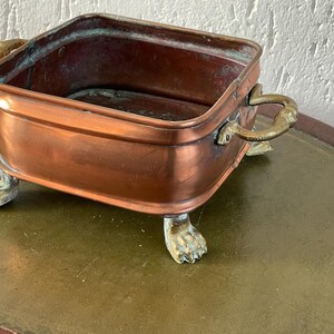 Puede incluir: Una jardinera cuadrada de color cobre con asas de latón y patas de garra. La jardinera tiene un aspecto ligeramente envejecido, con pátina en el cobre y el latón. Las asas son curvas y las patas están detalladas.