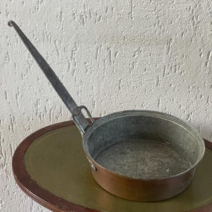 Könnte beinhalten: Eine antike Bratpfanne aus Kupfer und Metall mit einem langen, gebogenen Griff. Die Pfanne hat ein rundes, offenes Design mit einer Kupferaußenseite und einer Metallinnenseite. Der Griff ist aus dunklem Metall, und die Pfanne steht auf einem runden Tisch.