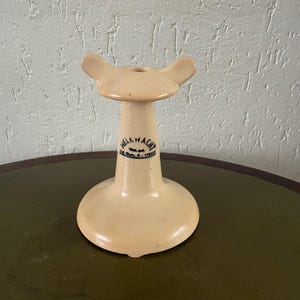 Op de afbeelding: Een crèmekleurige keramische standaard met een uitlopende basis, een centrale kolom en een bovenkant met twee vleugelachtige verlengingen. De standaard heeft een cirkelvormige opening aan de bovenkant en een zwart-blauw tekstlabel. De standaard is waarschijnlijk vintage.