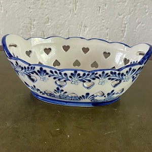 Op de afbeelding: Een witte keramische kom met een blauw bloemen- en hartontwerp. De kom heeft hartvormige uitsparingen rond de rand en een geschulpte rand. De kom heeft een diameter van ongeveer 25 cm.