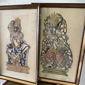 Op de afbeelding: Twee ingelijste schaduwpoppen, een met een vrouwelijke figuur in een blauw en gouden kostuum, de andere met een vrouwelijke figuur in een rood en gouden kostuum.