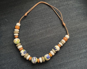 Puede incluir: Un collar de cordón de cuero marrón con cuentas de cerámica blancas y marrones. Las cuentas están decoradas con motivos florales azules y amarillos.