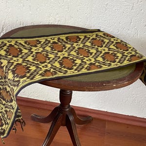 Può includere: Tessuto decorativo con un motivo ripetuto di forme floreali marroni e gialle, bordato con finiture nere. Il tessuto è drappeggiato su un tavolino rotondo in legno con piano verde scuro e base a tre gambe.