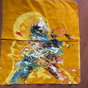 Può includere: Un quadrato di tessuto giallo con un design astratto colorato. Il design presenta un grande cerchio con spirali e un'esplosione di schizzi di vernice blu, verde e arancione.