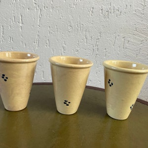 May include: Three ceramic cups with a light beige glaze and black polka dots. The cups are arranged in a row on a wooden surface.