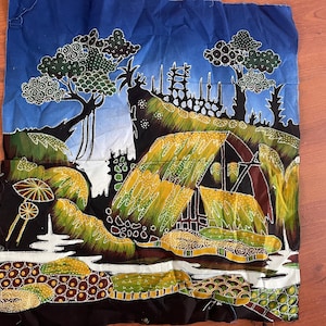 Op de afbeelding: Een kleurrijk batiktextiel met een landschapsscène met bomen, hutten en water. Het kunstwerk gebruikt een palet van blauw, groen, geel en bruin. Het ontwerp is gedetailleerd met ingewikkelde patronen en vormen, waardoor een levendige en gestructureerde uitstraling ontstaat.