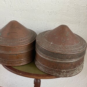 Op de afbeelding: Twee antieke koperen containers met ingewikkelde ontwerpen en een afgerond, conisch deksel. De containers zijn op elkaar gestapeld en hebben een versleten, verweerde patina.