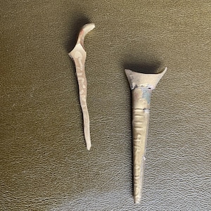 Op de afbeelding: Twee antieke metalen gereedschappen, één met een gebogen handvat en de andere met een conische vorm. De gereedschappen zijn zilverkleurig en lijken te zijn gemaakt van een metaallegering. De gereedschappen zijn geplaatst op een getextureerd, olijfgroen oppervlak.