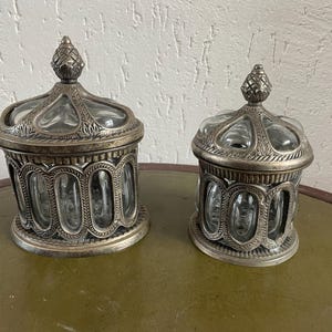 Może przedstawiać: Dwa ozdobne pojemniki wykonane ze srebra i szkła z ozdobnymi wzorami. Większy pojemnik znajduje się po lewej stronie, a mniejszy po prawej. Oba pojemniki mają przezroczyste szklane wnętrze i srebrne zewnętrzne z misternymi wzorami.