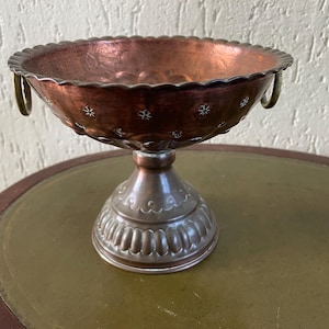 May include: A copper-coloured pedestal bowl with a scalloped rim and small star-shaped cutouts. The bowl has two small handles and sits on a decorative base. The bowl is likely vintage or antique.