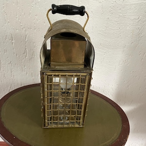 Op de afbeelding: Een messing lantaarn met een zwarte handgreep en een draadkooi. De lantaarn heeft een helder glazen paneel en een gloeilamp erin.