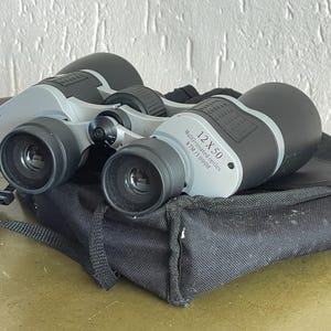 Op de afbeelding: Zilveren verrekijker met zwarte accenten en een zwarte draagtas. De verrekijker heeft de tekst "12X50 Multi-coated optics 87M / 1000m" erop gedrukt.