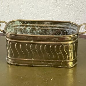 Peut inclure: Un pot rectangulaire en laiton avec un motif ondulé sur les côtés et des poignées décoratives. Le pot a un aspect vieilli, avec une oxydation verte visible. Le design général est vintage.