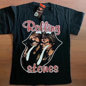 Puede incluir: Camiseta negra con el logo de los Rolling Stones, un diseño rojo y blanco con el nombre de la banda y un gráfico de los miembros de la banda. La camiseta tiene cuello redondo clásico y mangas cortas. Una etiqueta está adherida al cuello.