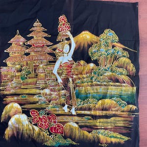 Op de afbeelding: Een zwarte wandhanger van stof met een kleurrijk batik-ontwerp. Het ontwerp toont een vrouw in traditionele Balinese kleding, een tempel en een berg. De kleuren zijn goud, rood, groen en blauw.