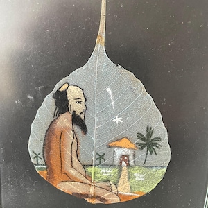May include: A hand-painted leaf art piece depicting a person sitting in a meditative pose with a small house and palm trees in the background. The leaf is translucent and the painting is done in shades of brown, green, and orange.