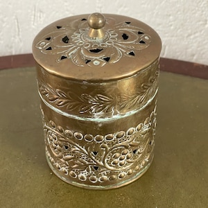 Peut inclure: Un récipient en laiton avec un design orné. Le récipient a un couvercle avec un bouton sur le dessus. Le récipient est décoré de motifs floraux et a une bordure perlée.