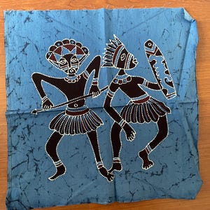 Puede incluir: Cuadrado de tela azul con un diseño batik que presenta dos figuras estilizadas en marrón oscuro y blanco. Las figuras están adornadas con tocados y faldas, sosteniendo lanzas y un escudo. El fondo tiene un patrón moteado.