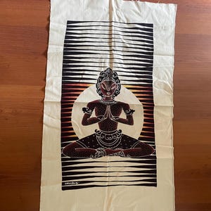 Op de afbeelding: Een witte wandhanger van stof met een zwart-wit gestreepte achtergrond. Een zwart-bruine illustratie van een persoon in meditatieve houding is gecentreerd op de stof. De persoon zit met gekruiste benen en de handen in gebedspositie. De illustratie is omgeven door een witte cirkel met een rood-oranje verloop.